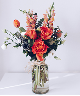 Vase filled with a vibrant arrangement of orange and white flowers including roses and tulips, set on a white table against a plain light background, creating a fresh and cheerful atmosphere